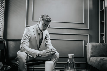 Valmiera, Latvia - September 9, 2023 - Thoughtful groom in a suit sitting in a stylish room, holding a glass, captured in a black and white photo before the wedding ceremony...