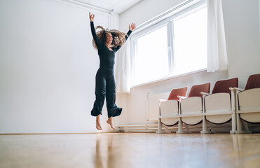 Fototapeta premium Woman in a black outfit gracefully jumping in a well-lit room, her movements fluid and expressive. Her curly hair flows as she extends her arms, embodying elegance and passion in her dance