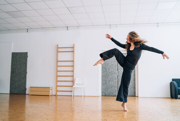 Woman in a black outfit gracefully dances in a well-lit room, her movements fluid and expressive. Her curly hair flows as she extends her arms, embodying elegance and passion in her dance