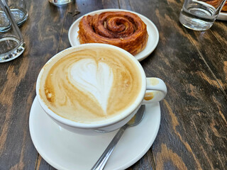 A cup of coffee with a heart design on it sits on a white plate next to a cinnamon roll