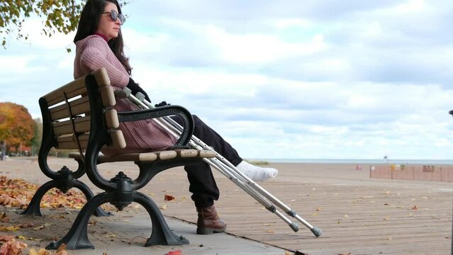 A woman sits on a bench with a cane in her hand. The bench is located on a pier overlooking the ocean. The woman is in a contemplative mood
