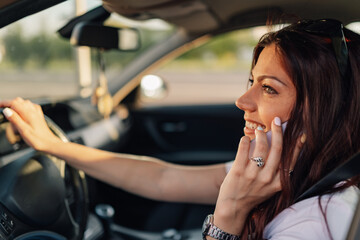 Trendy smiling woman driving a car and having a phone conversation.
