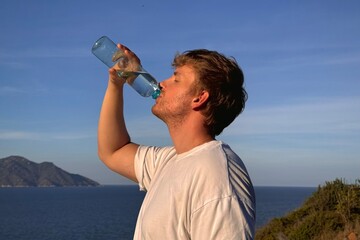 young man drinking water from bottle outdoors at sea, nature at summer hot day