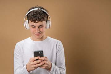One man caucasian young male stand studio shot on beige background use mobile phone smartphone with headphones listen music send messages texting or browse internet online app copy space
