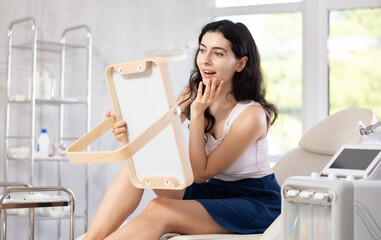 Young brunette woman admires her new face in the mirror after plastic surgery