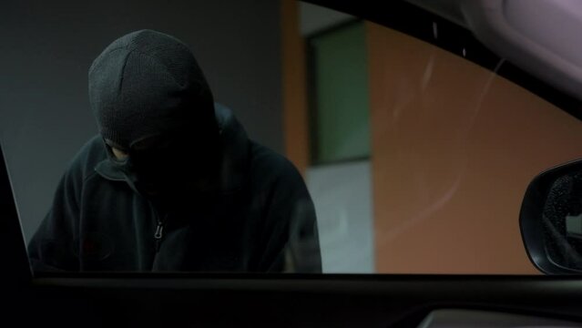 Inside view of the creepy thief with wearing black mask looking inside the car and try to break unlock the door's handle of vehicle, car security system to prevent crimination.
