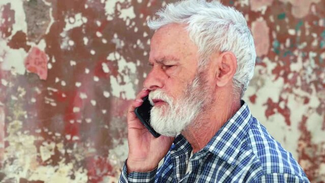 An elderly man with white hair and a white beard, wearing a blue and white checkered shirt, stands and talks on the phone. The background features a weathered, time-worn wall.
