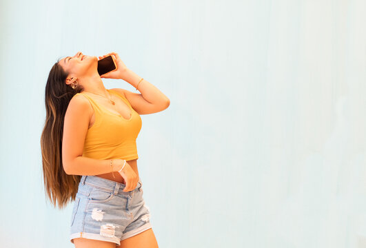 Mujer feliz con polera top amarillo posando aislada sobre fondo de pared gris hablando por tel&eacute;fono m&oacute;vil feliz, sonrisa,	
