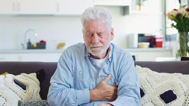 Worried elder senior man feeling bad, upset old middle aged grandfather touching chest feel sudden pain heartburn having heart attack sit on sofa at home
