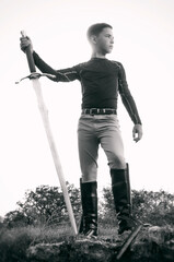 Dramatic black and white portrait of a teenage male standing on a rocky outcropping, holding a heavy iron sword at his side, as the evening sun sets behind him