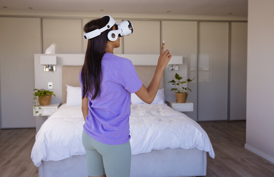 Happy biracial woman using virtual reality headset in modern bedroom