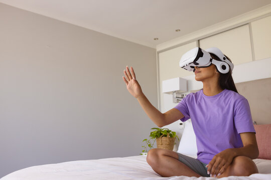 Happy biracial woman using virtual reality headset while sitting on bed, copy space