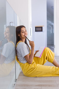 Happy biracial woman sitting on floor holding notebook and pen, smiling thoughtfully