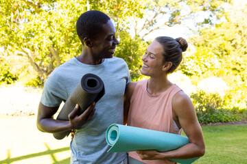 A diverse couple with yoga mats smile in a sunny park wellness and healthy lifestyle