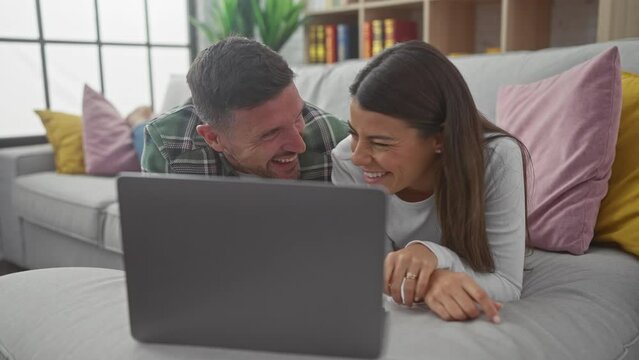 A happy couple lounging on a sofa indoors, smiling at something on a laptop.