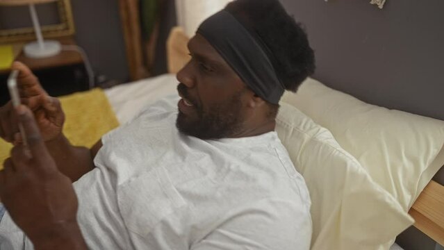 A handsome young african american man uses a smartphone while sitting in bed in a cozy bedroom at home, showcasing a comfortable indoor lifestyle.