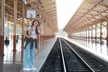 Young Female Traveler at Train Station Platform Ready for Adventure, Wearing Backpack and Hat, Smiling and Looking Excited for Journey, Sunlight Streaming Through Wooden Roof Structure