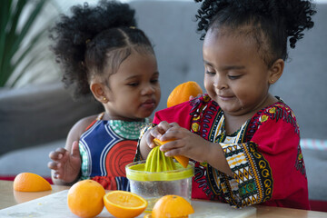 Two little African girls wearing traditional clothes have fun squeezing orange juice and trying to...
