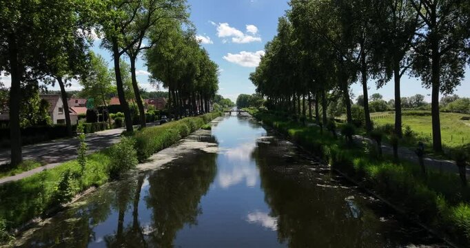 Damme, Belgium, June 2022. Drone pushes forwards above the Damse Vaart a peaceful canal lined with trees under a bright blue sky with fluffy clouds reflecting in calm water.