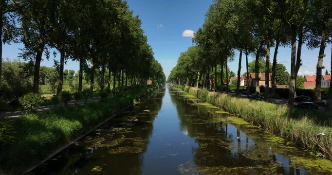 Damme, Belgium, June 2022. Drone pushes forwards above the Damse Vaart a canal lined with trees under a bright blue sky with fluffy clouds reflecting in calm water.