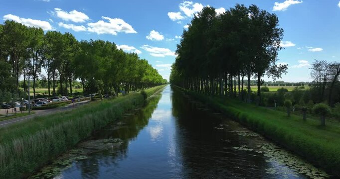 Damme, Belgium, June 2022. Drone pushes forwards above the Damse Vaart a serene canal lined with trees that stretch into the distance under a bright blue sky.