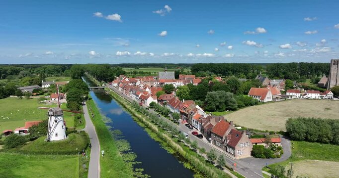 Damme, Belgium, June 2022. Drone orbits away from the Schellemolen windmill towards the town with historic buildings, showcasing the Damse Vaart 
canal and lush greenery on a sunny day.