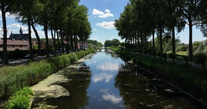 Damme, Belgium, June 2022. Drone pushes forwards above the Damse Vaart a picturesque canal with lush greenery under a bright blue sky with fluffy clouds.
