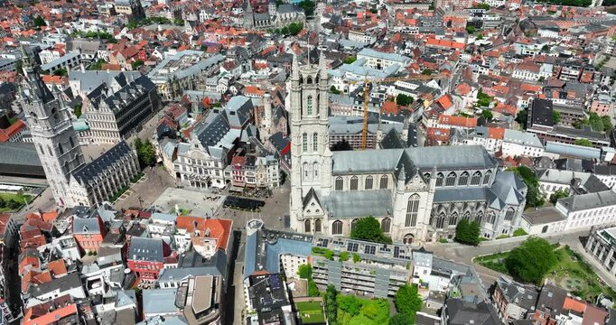 Saint Bavo's Cathedral, East Flanders, Ghent, Flemish Region, Belgium, June 2022. Drone orbits right as sunlight illuminates town and tall tower facade with cheerful lighting.