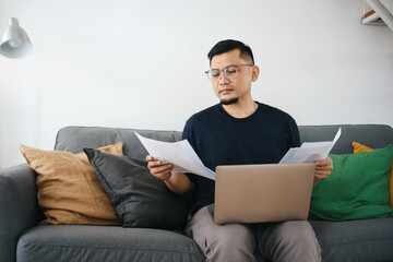 Focused Asian man sitting on couch looking at paper documents and using laptop. Work from home concept.