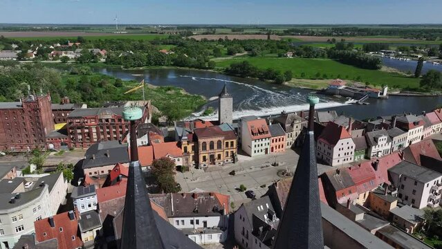 Flight towards the weir in the river Saale in Calbe. Movement through the church towers of the St. Stefani church. Town hall and town hall square below. Travel destination in Saxony-Anhalt.