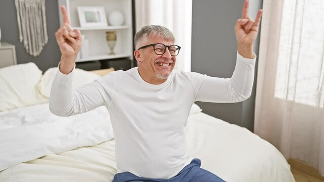 Madcap middle age grey-haired man rocking out in pyjamas, striking wild heavy music star expression, throwing up the rock hand symbol from his bedroom.