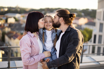 Mother and father kissing daughter on rooftop at sunset. Displaying love, togetherness, and family bond. Warm daylight.