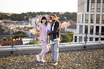 Fototapeta premium Cheerful family with parents and daughter posing for selfie on rooftop with cityscape in background during sunset.
