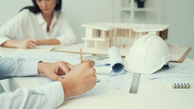 Professional architect engineer drawing blueprint and working together with young beautiful caucasian coworker on meeting table with safety helmet, house model, blueprint scatter around. Immaculate.