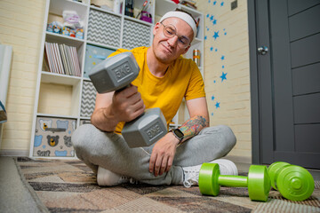 A man in a yellow shirt and grey sweatpants sits on the floor lifting a grey dumbbell. He is in a...