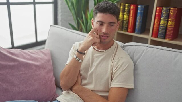 Pensive, young hispanic man sits on living room sofa, finger to chin, deep in concentrated thought, wrestling with doubt, lost in a world of wonder at home.