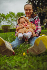 A mother and her baby enjoy a beautiful spring day in the park. They are sitting on a blanket, surrounded by lush green grass and blooming flowers.