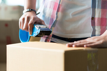 Concept young couple moving house. Close-up hand of man use tape sealing cardboard box.