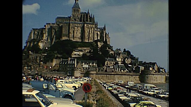 Mont Saint-Michel, FRANCE - July 12,1976: ground panorama with tourists in the car parking of Historic Saint Michael's Mount in the 70s. People in vintage 1970s dress and vintage cars and bus.