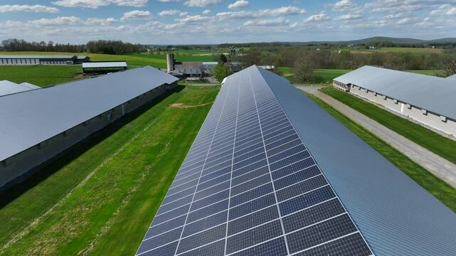 Modern farm with solar panels installed on roof of barn building. Sunny day in rural countryside of USA. Drone flyover shot.
