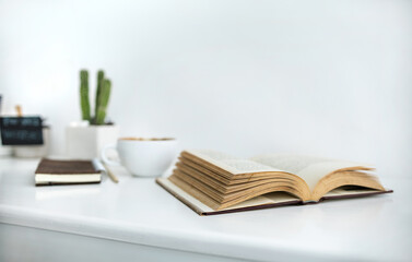 Close-up shot, open book with white coffee cup and note book on white table and white wall on background