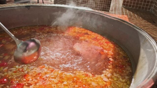 Chef mixes meat soup with vegetables in a large cauldron with a slotted spoon