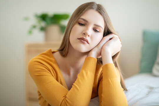 A teen girl, dressed in a yellow shirt, sits indoors with a thoughtful expression on her face. The background is softly lit, and a potted plant is visible behind her