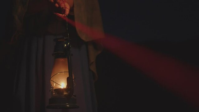 A woman in vintage clothes in the dark with her hands tied with a red thread holds a kerosene lamp. Soft light on hands. Psychology