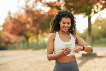 A young woman wearing sports attire is standing in a park during an autumn afternoon. She is looking at her fitness tracker while holding a smartphone, surrounded by colorful fall foliage.