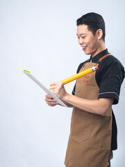 Smiling young man in a black uniform and brown apron, holding a large novelty pencil and a white clipboard against a plain light gray background