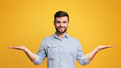 Smiling millennial man holding empty palms up as scales, making choice, copy space, yellow studio background