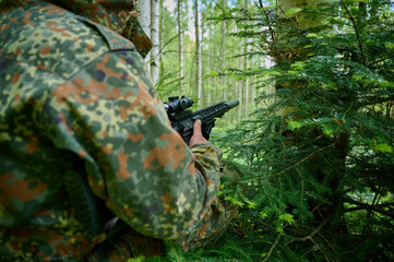 Hunter with rifle wearing camouflage suit sitting in ambush. Camouflaged sniper in the forest in ambush. Military man hide in the woods with rifle.