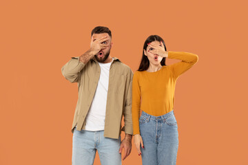 Obraz premium A young couple stands side by side against an orange backdrop, each covering one eye with their hand and looking surprised.