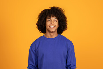A young man with curly hair is smiling happily while standing indoors. He is wearing a blue...